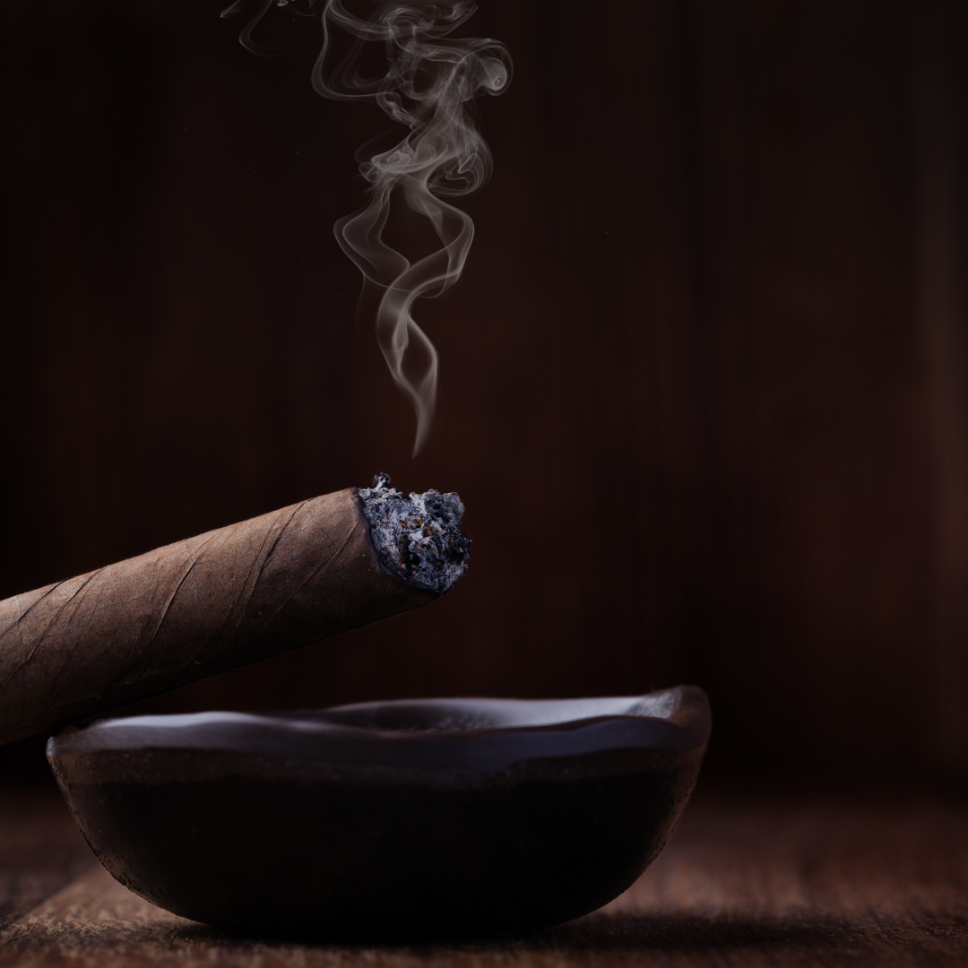 Cigar with ash resting on a dark ashtray against a dark background