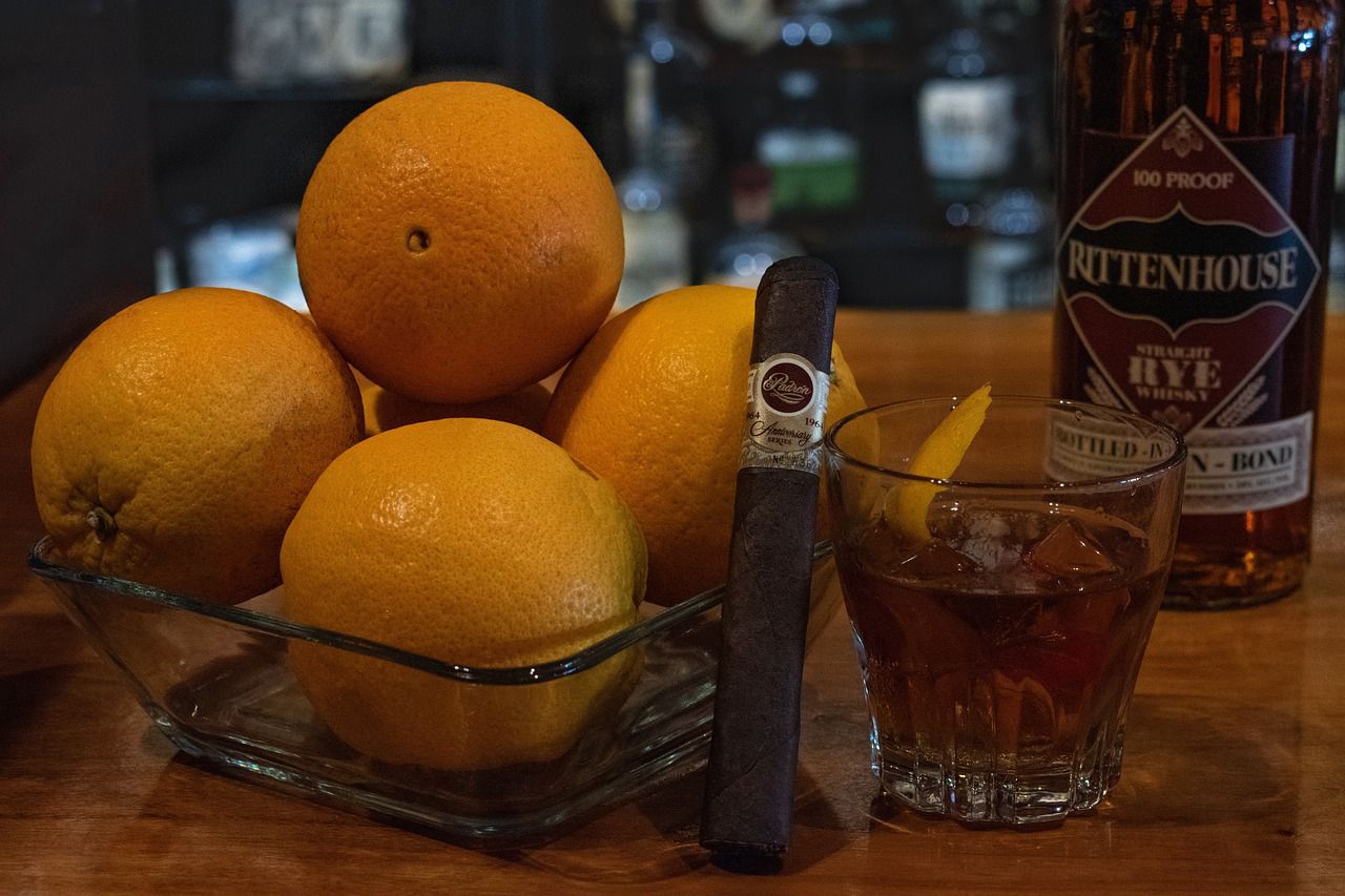 Oranges in a glass bowl with a glass of rye and a cigar on a wooden surface, next to a bottle of Rittenhouse Rye.